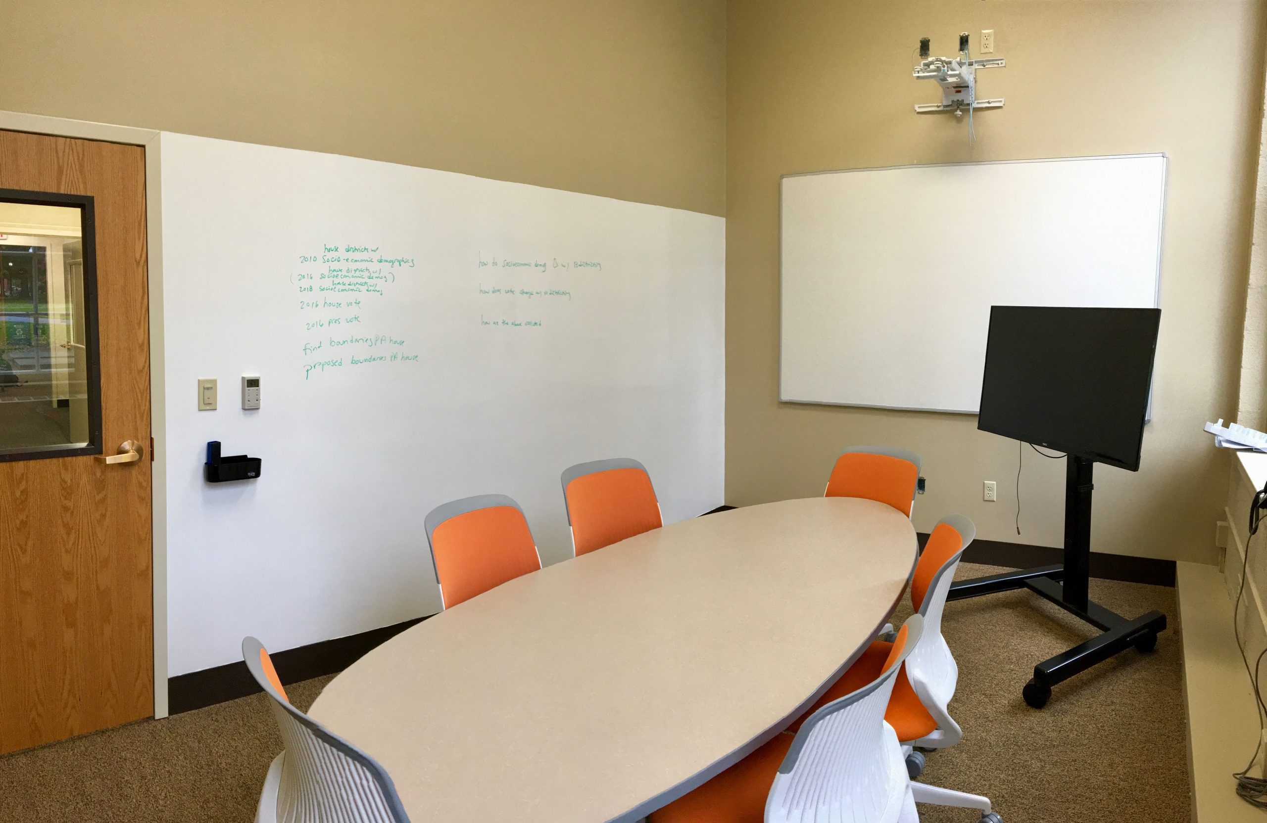 A study room with a long table, six chairs, white boards, and a portable screen