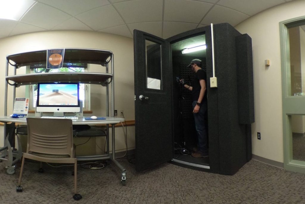 A person standing in a small recording booth next to a desk with a Mac computer