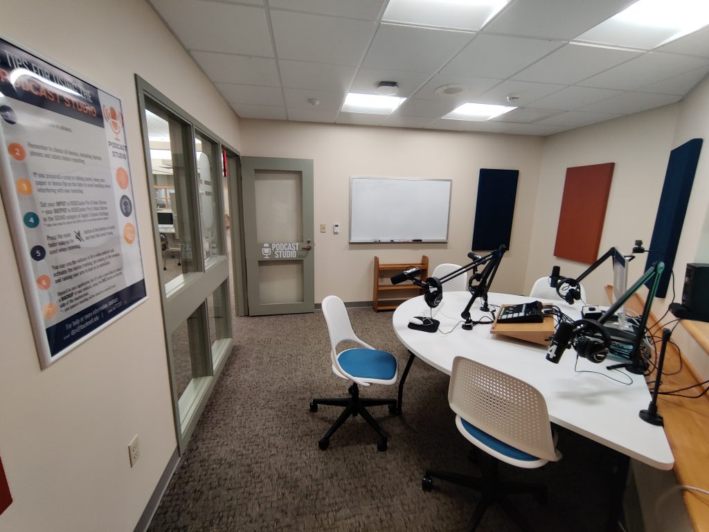 The Podcast Studio, a room with sound panels on the walls, a large "Tips for using the Podcast Studio" poster, and a half-circle table with four chair around it, mounted microphones, a switchboard, and a Mac computer.