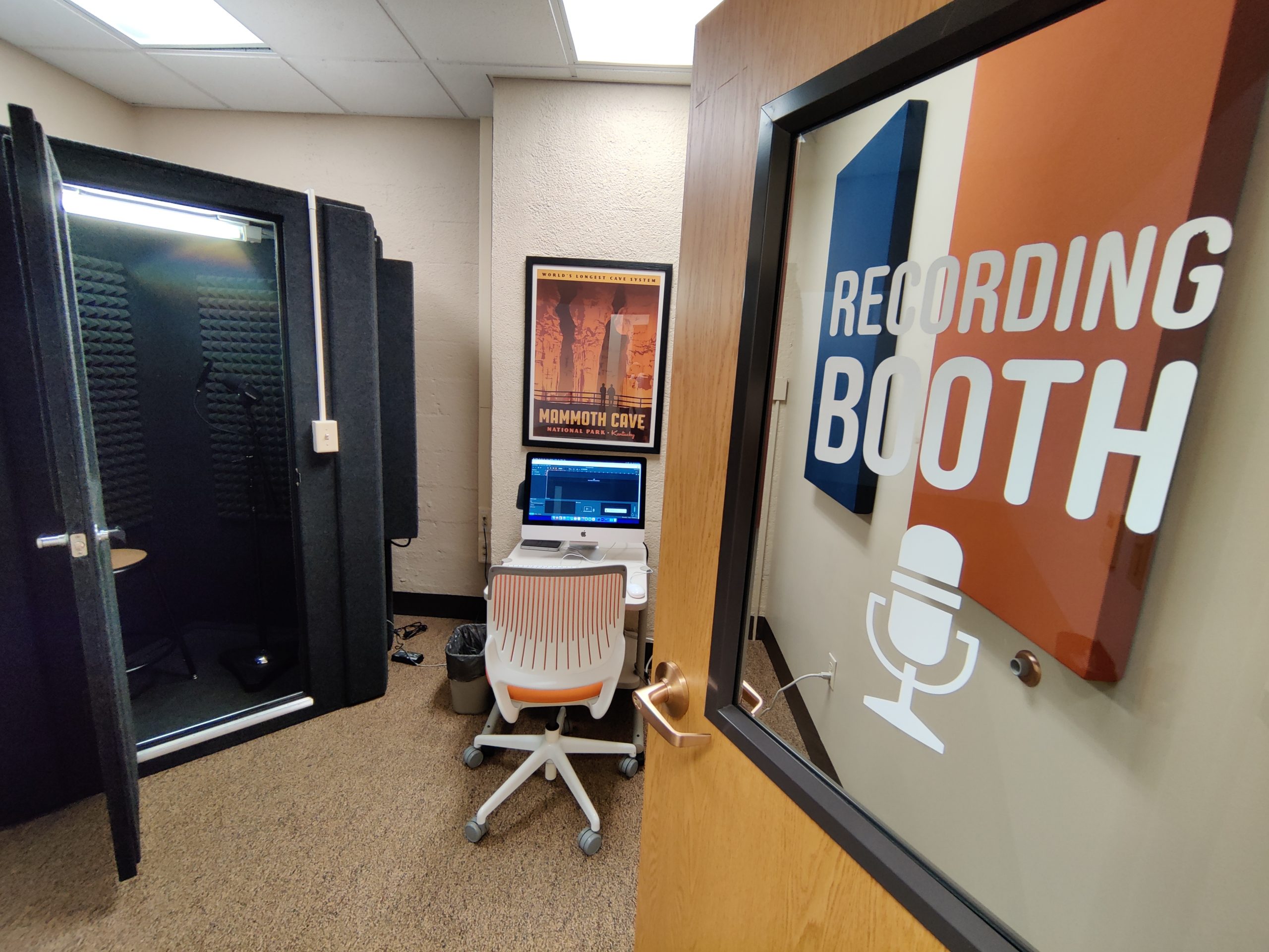 The interior of the Recording Booth room, showing a small room with no windows containing a table, rolling chair, a Mac, and a full recording booth.