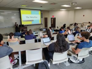 Kate Tuley teaching at the front of a classroom full of students in front of a slide that reads, "The MetaZine."