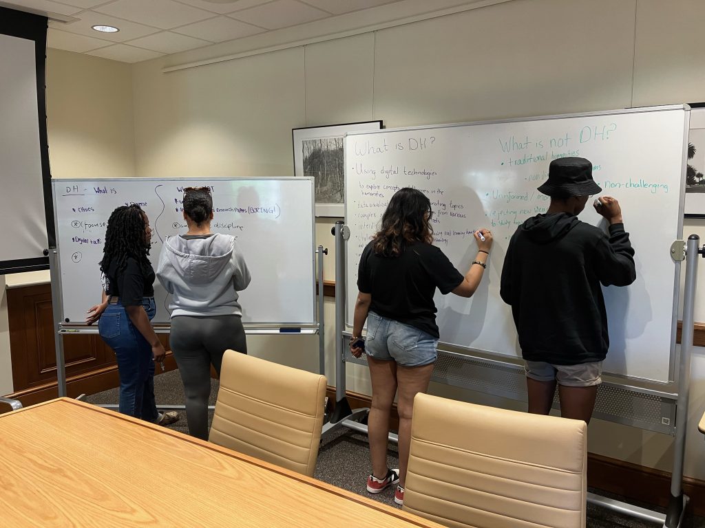 Jackline, Gianna, Malika, and Thandeka writing on two whiteboards under the headings "what is DH?" and "what is not DH?"