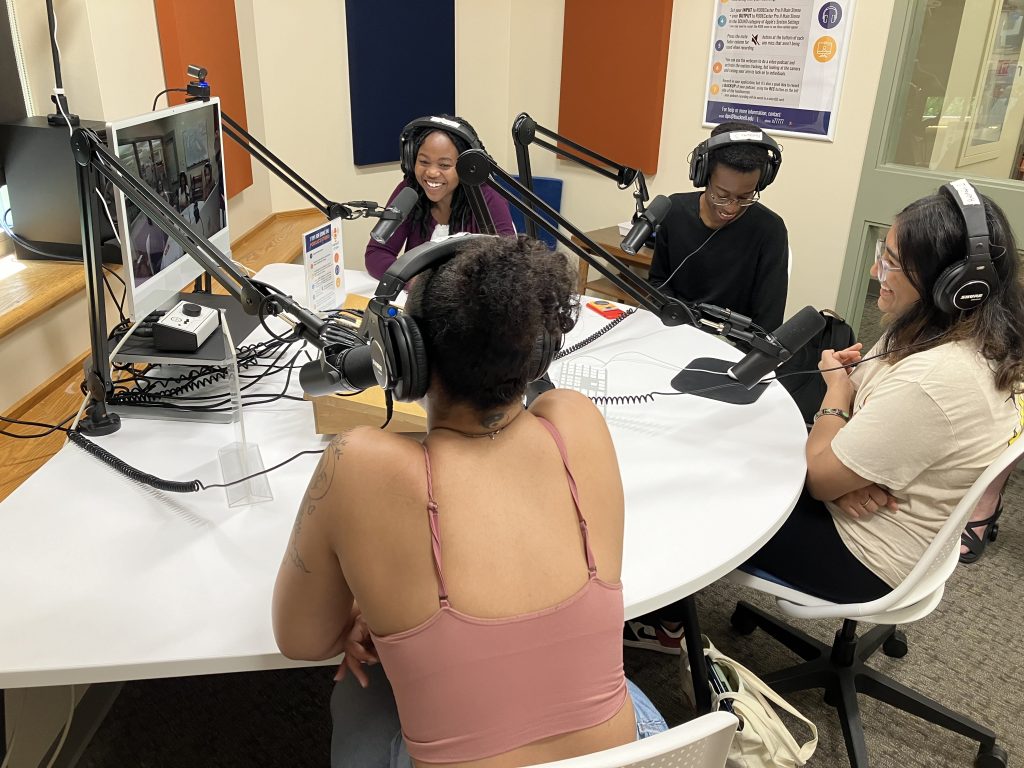 Gianna, Malika, Thandeka, and Jackline all sitting in front of microphones, talking and laughing in the Bertrand Library Podcast Studio.