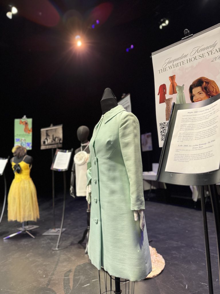 a pale turquoise jacket draped upon a stand in the forefront of a presentation flanked by stands with paper descriptions, a large poster decribing the Jacqueline Kennedy White House years,  and additional blurred dresses on a stage in the background.